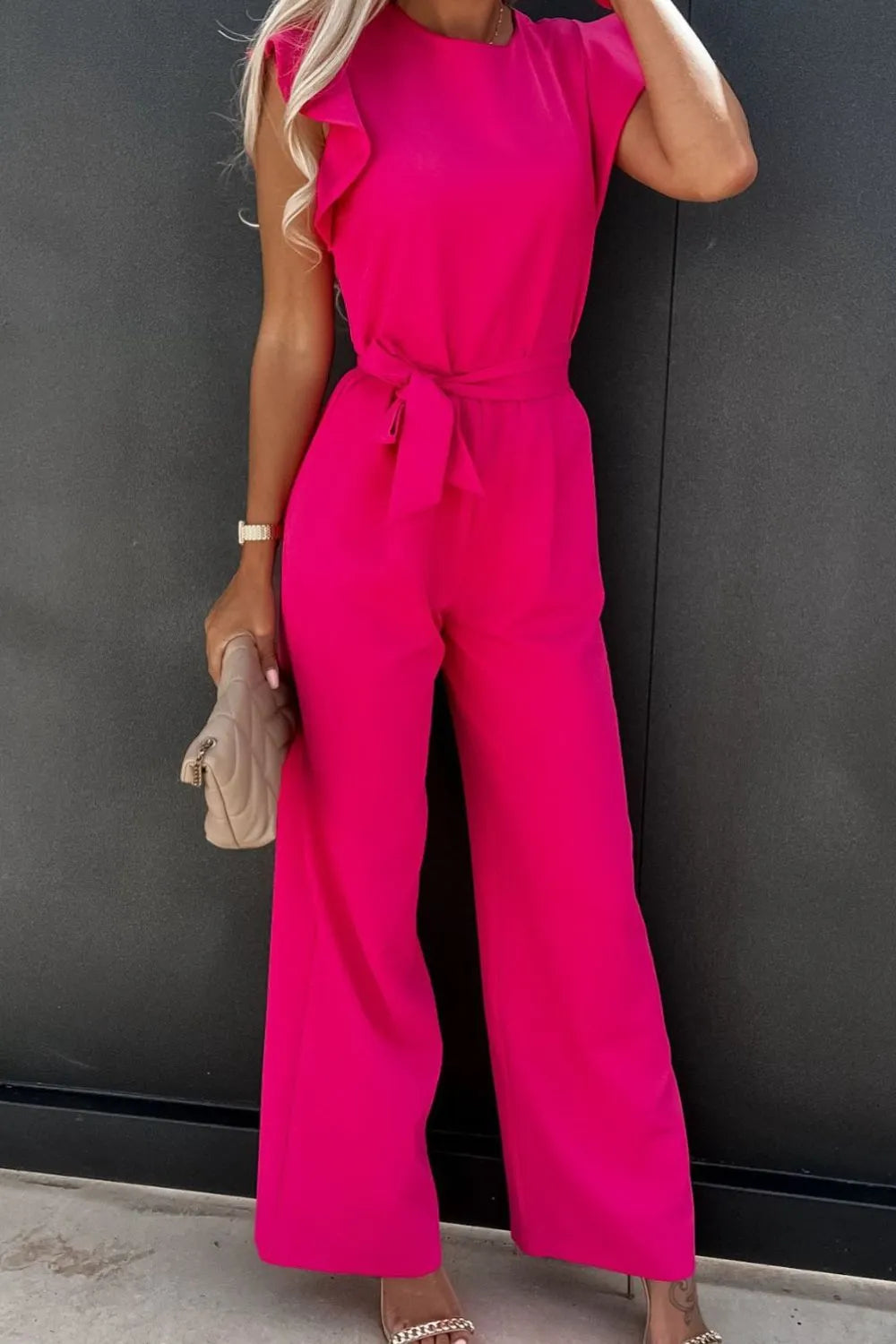 Model wearing a bright pink round neck cap sleeve jumpsuit with a tie waist and ruffled sleeves.