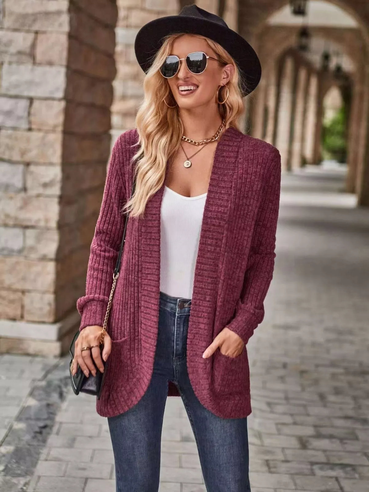 Stylish woman in open front ribbed knit cardigan, sunglasses, and hat, walking in a stone archway.
