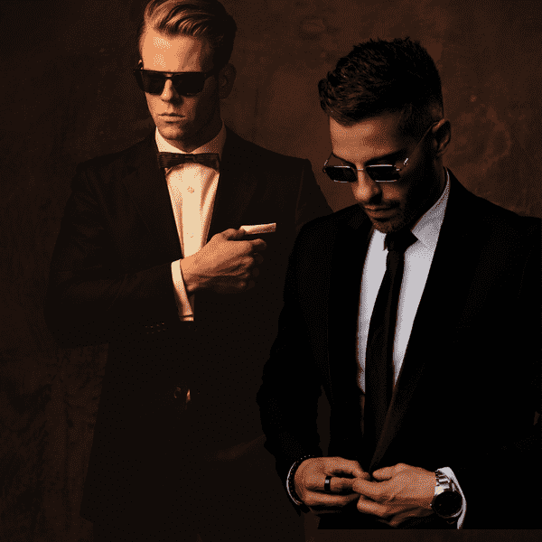 Two men in suits and sunglasses posing against a dark background