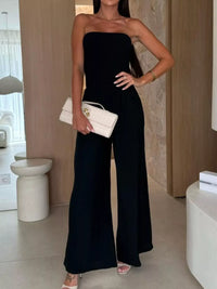 Woman in a black smocked tube wide-leg jumpsuit holding a cream handbag, styled for formal wear in NZ.